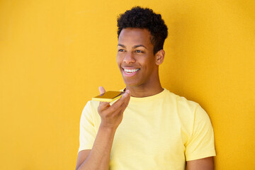 Smiling man making voice note through smart phone in front of yellow wall