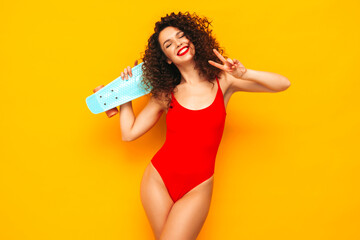 Young beautiful sexy smiling woman posing near yellow wall in studio.Trendy model in red summer swimwear bathing suit.Positive female with afro curls. Happy and cheerful. Holding penny skateboard