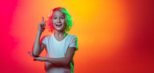 Half-length portrait of little boy, cute kid raising finger up isolated on mangenta yellow studio background in neon light, filter.