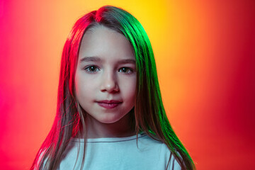Close-up preschool girl, calm kid with long glossy hair looking at camera isolated on red yellow studio background in neon light, filter.