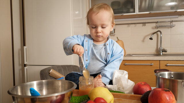 Little baby boy sitting on kitchen table and eating fruit puree with spoon. Concept of healthy nutrition,diet and children food