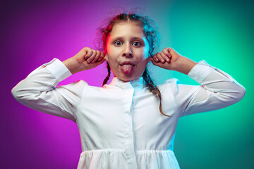 Half-length portrait of funny little girl, kid making faces isolated on gradient purple blue color background in neon light