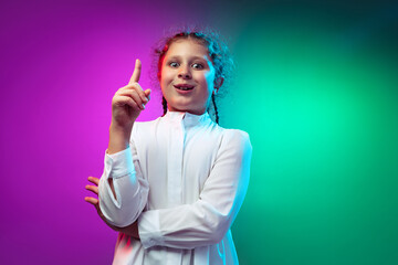 Portrait of little preschool girl, cute kid raising finger up isolated on blue purple studio background in neon light, filter.