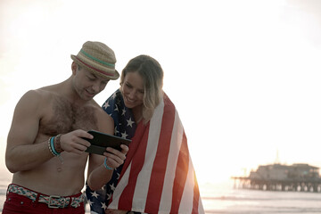 Handsome man using digital tablet by girlfriend wrapped in flag on Santa Monica beach