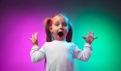 Studio shot of surprised little girl, kid standing with open mouth isolated on gradient purple blue color background in neon light