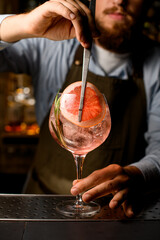 Close-up view of goblet glass with cold cocktail that the hand of barman decorating by a slice of grapefruit using tweezers
