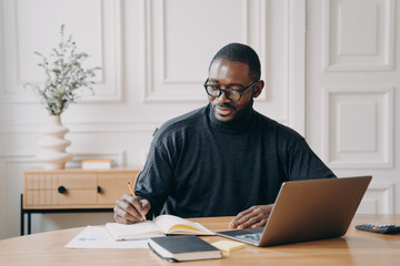 Focused young african american businessman writing down notes in notebook while working in office