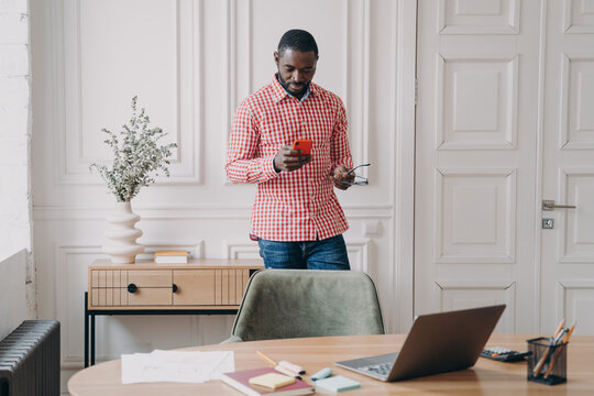 Young serious african american businessman in casual clothes standing in office with smartphone