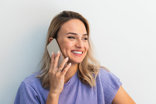 Smiling young woman looking away while talking on mobile phone in front of wall
