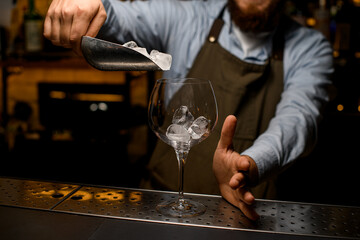 transparent wine goblet glass in which bartender pours ice cubes from a scoop