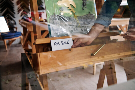 Male store owner keeping for sale sign in front of painting seen through glass window