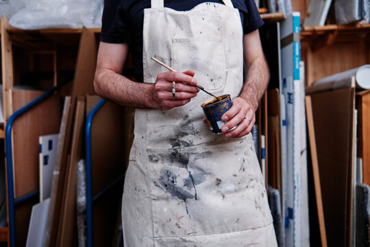 Male frame maker wearing apron holding paint brush and can while standing in workshop