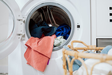 Close-up of colorful clothes in a basket, in the background stands an open washing machine with laundry thrown in, weekend cleaning, household chores