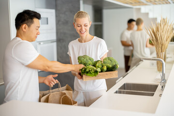 Naklejka premium Asian man with shopping bags and woman with fresh vegetables on kitchen at home. Multiracial couple ready to cooking and look at camera. Concept of healthy and vegetarian eating. Idea of shopping