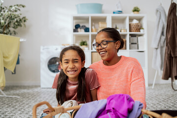 Smiling woman wearing glasses is sitting with pretty daughter on floor of bathroom laundry room in front of them wicker basket with colorful clothes in background dryer hangers washing machine