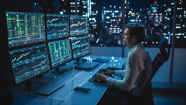 Financial Analyst Working on a Computer with Multi-Monitor Workstation with Real-Time Stocks, Commodities and Foreign Exchange Charts. Businessman Works in Investment Bank City Office Late Evening.