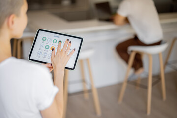 Woman pushing button on kitchen app of smart home on digital tablet at home. Concept of modern home control. Idea of domestic lifestyle. Cropped image of girl. Blurred man on background using laptop