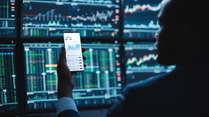 Financial Analyst Using Smartphone with Real-Time Stocks, Commodities and Market Charts. Working on Multi-Monitor Workstation with Stock Charts. Businessman Works in Investment Bank in the Evening.