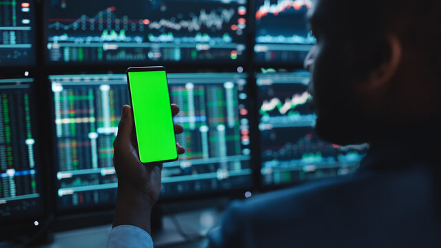 Financial Analyst Using Smartphone with Green Screen Chroma Key Mock Up Template and Working on Multi-Monitor Workstation with Stocks Charts. Businessman Works in Investment Bank in the Evening.