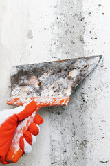 Hand of plasterer holds an old flexible blade spackle knife and scrapes concrete wall, removes plaster during redecoration of building. Close-up view, selective focus on working tool putty knife.