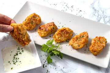 Hand holding a vegetarian nugget dipping it in a yogurt sauce with parsley leaves on white marble background