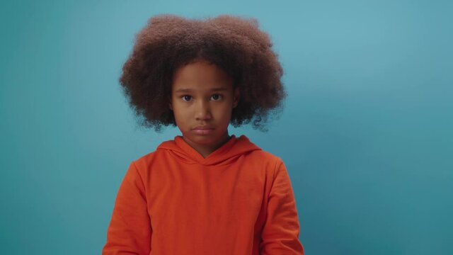 Upset African American girl looking at camera standing on blue background. Kid shows unhappy emotion. 