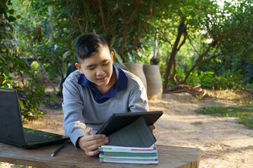 The boy is studying online on a wooden table with a laptop computer and tablet during morning hours...
