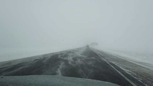 Steadicam shot of car driving on a siberian intercity highway when strong wind and heavy snowstorm in evening. Some cars and driving ahead. Fields on both sides of the road