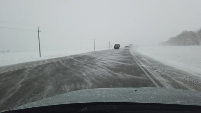 Steadicam shot of driving on a siberian highway when strong wind and heavy snowstorm in evening. Some trucks driving in the oncoming lane with headlights on. Field is on left side of the road