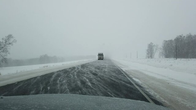 Steadicam shot of car driving on a siberian highway when strong wind and heavy snowstorm in evening. Cars and truck driving in the oncoming lane with headlights on. Fields and trees near the road