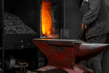 Empty red anvil in the forge. A blacksmith kindles a flame in the forge in the background.