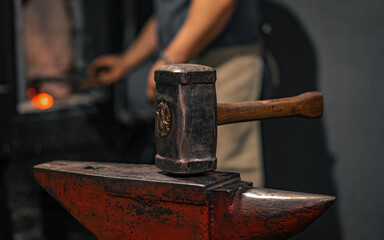 The blacksmith's hammer is on the anvil in the forge. A blacksmith lights a forge in the background.