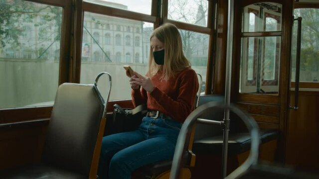 Cinematic shot of young woman in face mask during corona virus regulations, travel in tram or bus, use smartphone to check news. Woman in public transport during 2022 corona rules