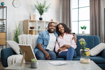 Beautiful and happy couple sitting on couch in embrace and smiling on camera. African american man...