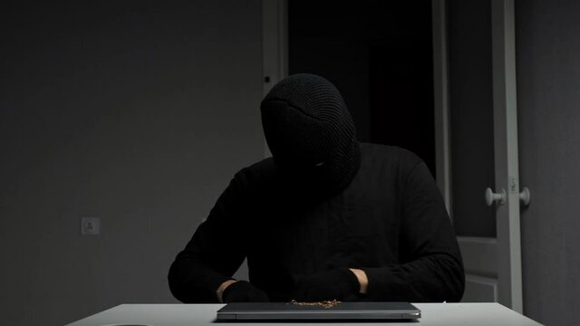 Happy with godsend thief in black mask and gloves counts money cash and throws up stolen banknotes and puts jewelry on laptop sitting at wooden desk, closeup.