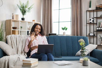 Positive african american woman sitting on couch, smiling and waving hand during video chat on modern laptop. Concept of people, technology and communication.