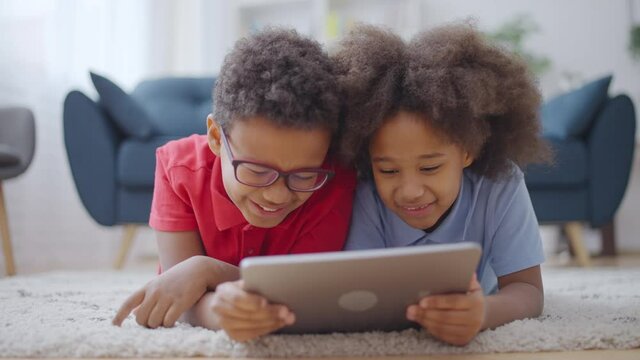 Cheerful black siblings lying on floor with tablet, having fun together, smiling