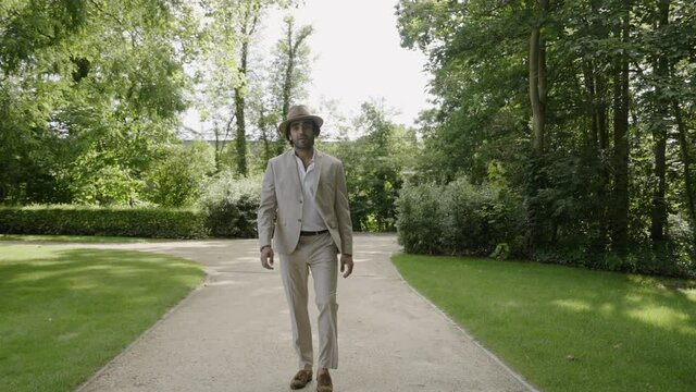 Confident, stylish man wearing a suit and hat walking through a garden towards the camera and taking off his hat, concept for luxury lifestyle, success and wedding, 4K 120FPS 10bit slowmotion