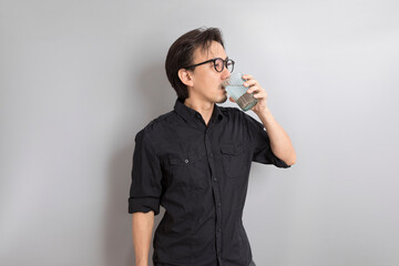 Asian man with glasses of water wearing black shirt drink fresh water stand on grey background.