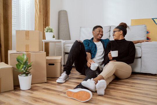 Smiling couple is sitting on floor relaxing after moving in, drinking coffee in their new apartment, around them boxes with unpacked things, husband embraces his wife and looks at each other happily