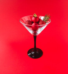 The concept of celebrating the New Year. A martini glass with shiny red Christmas balls and a sprig of spruce on a red background.