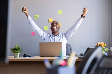 Cheerful businessman with arms raised while sitting with laptop at office