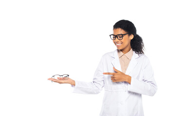 Smiling african american doctor pointing at eyeglasses on hand isolated on white.