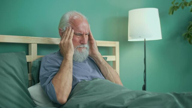 Tired Elderly Man Has Acute Headache While Sitting on ahe Bed At Home. Virus, Headache, Migraine, Anxiety. Seniors, Dizziness, High Blood Pressure, Health Care and Retirement Insurance.