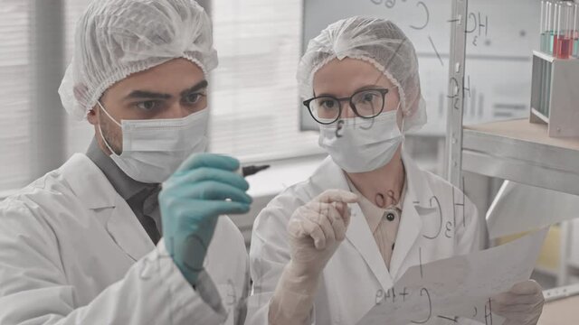 High-angle of Biracial man writing structural chemical formulas on glass wall in laboratory, female Caucasian colleague standing near and holding paper, scientists wearing medical gowns and masks