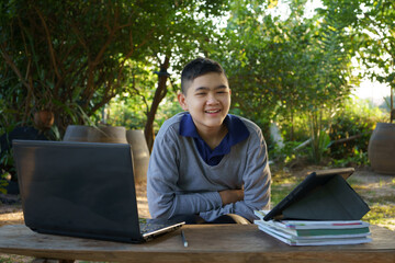 Boy looking at camera studying online on a wooden table with laptop computer and tablet in the morning in a rural home. concept online education countryside area and work from home