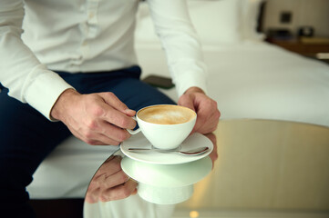 Close-up of a white ceramic cup with delicious coffee drink, cappuccino in the hands of an unrecognizable man in business casual attire, sitting on the bed in hotel room