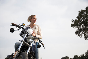 Beautiful female biker holding helmet on motorcycle while looking away in front of sky