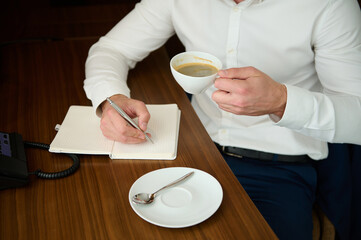 Cropped view of unrecognizable entrepreneur holding a cup with freshly brewed coffee and makes notes on a notepad, writing text, sitting at desk in hotel room during business trip
