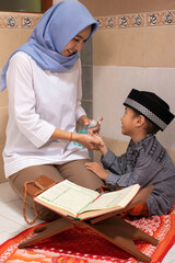 a Muslim Asian mother is wearing a sanitizer to her son who is reading the Al-Qur'an